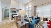 This thoughtfully designed single-floor residence showcases a contemporary aesthetic with impressive soaring ceilings and an open-concept layout that seamlessly connects the living and kitchen areas. The kitchen is a functional centerpiece, featuring warm wood-tone cabinetry, white quartz countertops, stainless steel appliances, a tile backsplash, and a peninsula island with bar seating. Clerestory windows flood the space with abundant natural light, complementing the light hardwood flooring that extends throughout the main living areas. The living room offers a welcoming atmosphere anchored by bold, vibrant furnishings and large windows dressed with blinds and patterned curtains. Two well-appointed bedrooms are carpeted for comfort, each offering generous proportions and natural light. The primary bedroom features vaulted ceilings and a spacious walk-in closet with built-in shelving and hanging rods. Both bathrooms are finished with wood-tone vanities, white quartz countertops, large mirrors, and hardwood-style flooring. One bathroom includes a soaking tub, while the other features a shower. Throughout the residence, consistent modern finishes and a cohesive blue, white, and natural wood palette create a polished and inviting ambiance.