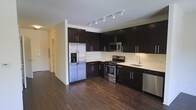 This single-floor residence offers a thoughtfully designed layout with a contemporary aesthetic that blends style and functionality. The kitchen serves as a standout feature, showcasing floor-to-ceiling dark espresso cabinetry that provides generous storage and a bold visual contrast against the crisp light countertops. A full suite of stainless steel appliances — including a French door refrigerator with ice and water dispenser, a gas range, built-in microwave, dishwasher, and a sleek undermount sink — equips the space for everyday cooking and entertaining alike. Track lighting runs along the ceiling, offering focused illumination throughout the kitchen area. Rich, dark hardwood flooring extends seamlessly from the kitchen into the adjacent living areas, lending warmth and cohesion to the open-concept floor plan. A hallway visible from the main living space suggests additional rooms beyond, contributing to a sense of depth and privacy within the layout. The overall ambiance is clean, modern, and move-in ready, with a neutral palette and quality finishes that provide a versatile backdrop for a variety of interior styles.