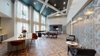 Located in Houston, TX 77008, this club room is a thoughtfully designed communal space that blends modern elegance with functional versatility. The room's most striking architectural feature is its dramatic double-height ceiling, accented by a deep teal coffered detail and recessed lighting that adds depth and visual interest overhead. Floor-to-ceiling windows line one wall, inviting an abundance of natural light and offering open views of the surrounding outdoor area, dressed with sheer white drapery for a refined finish. The geometric patterned hardwood flooring serves as a unifying design element throughout, adding warmth and a sophisticated visual rhythm to the expansive open layout. A large communal wood table with upholstered seating anchors the central gathering area, ideal for collaborative work or group dining. Adjacent to this, a comfortable lounge area features a generous sectional sofa centered around a linear fireplace set into a dramatic full-height stone accent wall, with a wall-mounted TV above. Built-in bookshelves and curated décor add a residential warmth to the space. A wet bar area with white quartz countertops, stainless steel appliances, decorative tile backsplash, and bar seating provides a convenient amenity for entertaining. Together, these elements create a polished, welcoming environment suited for a wide range of social and professional gatherings.