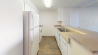 This single-floor space includes a well-organized galley-style kitchen that serves as a central feature. The kitchen is designed for efficiency, with cabinetry and countertops running along both sides of the room, maximizing storage and workspace. White upper and lower cabinets provide ample storage, while the light-toned laminate countertops offer a clean, neutral aesthetic that complements the overall color palette of the space.

A full suite of white appliances is included, featuring a refrigerator, electric range with oven, and a dishwasher, making the kitchen fully equipped for everyday use. The double-basin stainless steel sink, positioned along one counter run, adds a practical element to the layout. Overhead, a flush-mounted fluorescent light fixture provides consistent, even illumination throughout the kitchen.

The hardwood-style flooring runs the length of the kitchen, offering both durability and a warm visual contrast to the white cabinetry and neutral walls. The crisp white walls and bright overhead lighting contribute to an open, airy feel. The overall design prioritizes functionality and clean aesthetics, creating a practical and inviting space.