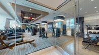 This well-appointed fitness center is designed to deliver a premium workout experience within a thoughtfully planned, single-level layout. The space is enclosed by floor-to-ceiling glass walls that provide an open, transparent feel while visually connecting different zones of the facility. Large windows line the perimeter, flooding the interior with natural light and offering views of the surrounding urban environment.

The cardio zone features a row of modern treadmills and stationary bikes positioned along the windowed wall, maximizing the outdoor sightlines for users during their workouts. Each machine is equipped with individual digital screens for an enhanced exercise experience. The strength training area is equally well-equipped, housing a multi-station cable and weight machine circuit, a power rack, adjustable benches, a full dumbbell rack, and kettlebells. Floor-to-ceiling mirrors line one wall of the free weights area, enhancing the sense of depth and allowing for proper form monitoring.

Overhead, large drum-style pendant light fixtures complement the recessed ceiling lighting, contributing to a polished, modern aesthetic. The flooring combines durable carpet tile with wood-tone sections. A water station is also available within the space, adding convenience for users.