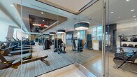 This well-appointed fitness center is designed to deliver a premium workout experience within a thoughtfully planned, single-level layout. The space is enclosed by floor-to-ceiling glass walls that provide an open, transparent feel while visually connecting different zones of the facility. Large windows line the perimeter, flooding the interior with natural light and offering views of the surrounding urban environment.

The cardio zone features a row of modern treadmills and stationary bikes positioned along the windowed wall, maximizing the outdoor sightlines for users during their workouts. Each machine is equipped with individual digital screens for an enhanced exercise experience. The strength training area is equally well-equipped, housing a multi-station cable and weight machine circuit, a power rack, adjustable benches, a full dumbbell rack, and kettlebells. Floor-to-ceiling mirrors line one wall of the free weights area, enhancing the sense of depth and allowing for proper form monitoring.

Overhead, large drum-style pendant light fixtures complement the recessed ceiling lighting, contributing to a polished, modern aesthetic. The flooring combines durable carpet tile with wood-tone sections. A water station is also available within the space, adding convenience for users.