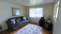 This two-floor home offers a straightforward and functional layout across its levels. The living room, visible in the provided image, features warm hardwood flooring that adds a classic, inviting character to the space. A large picture window dressed with vertical blinds allows natural light to fill the room while providing a view of the surrounding greenery outdoors. The neutral-toned walls create a versatile backdrop, complemented by a wall-mounted mirror that enhances the sense of openness. The room accommodates comfortable seating and additional storage or display furniture, making it a practical and livable space. A decorative area rug with a blue floral pattern anchors the seating area, adding a touch of visual interest to the room. The overall ambiance of the living area is calm and understated, with a layout that prioritizes functionality. The home's two-floor structure suggests additional living, sleeping, or utility spaces on the upper level, providing ample room for everyday living. Overall, this home presents a clean and livable environment with classic interior details.
