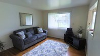 This two-floor home offers a straightforward and functional layout across its levels. The living room, visible in the provided image, features warm hardwood flooring that adds a classic, inviting character to the space. A large picture window dressed with vertical blinds allows natural light to fill the room while providing a view of the surrounding greenery outdoors. The neutral-toned walls create a versatile backdrop, complemented by a wall-mounted mirror that enhances the sense of openness. The room accommodates comfortable seating and additional storage or display furniture, making it a practical and livable space. A decorative area rug with a blue floral pattern anchors the seating area, adding a touch of visual interest to the room. The overall ambiance of the living area is calm and understated, with a layout that prioritizes functionality. The home's two-floor structure suggests additional living, sleeping, or utility spaces on the upper level, providing ample room for everyday living. Overall, this home presents a clean and livable environment with classic interior details.