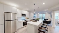 This thoughtfully designed single-floor residence showcases a clean, contemporary aesthetic with a cohesive neutral palette and light hardwood flooring throughout. The open-concept kitchen is a standout feature, equipped with stainless steel appliances including a full-size refrigerator, range, and built-in microwave. Flat-front white cabinetry extends to the ceiling for generous storage, while a large white quartz island with an undermount sink and bar seating serves as both a functional workspace and casual dining area. Stylish pendant lighting accentuates the island, seamlessly connecting the kitchen to the living area. The living room is bright and welcoming, benefiting from expansive sliding glass doors that flood the space with natural light and open to an outdoor balcony with open views. The bedroom is a serene retreat, featuring a large window with outdoor views, a wall-mounted TV, and a sliding door closet, with access to an organized walk-in storage area. The bathroom presents a refined, spa-like atmosphere with large-format marble-look tile flooring, a dark wood floating vanity with a white countertop, and a glass-enclosed walk-in shower finished in dramatic dark tile. Recessed lighting throughout maintains a polished, modern ambiance.