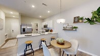 This thoughtfully designed single-floor residence offers a cohesive blend of modern finishes and warm, inviting aesthetics throughout. The open-concept kitchen is a standout feature, equipped with stainless steel appliances including a French door refrigerator, a built-in microwave, and an electric range. White shaker-style cabinetry pairs cleanly with light quartz countertops and a peninsula that doubles as a breakfast bar with stylish industrial-style bar stools. A pendant light fixture illuminates the adjacent dining nook, which comfortably fits a round table and chairs. The living room is generously proportioned with wood-look luxury vinyl flooring that runs throughout the unit, neutral wall tones, and ample space for relaxed seating arrangements. A wide entryway hallway connects the living areas to the private spaces, including a well-sized bedroom that benefits from large windows with outdoor views and abundant natural light. The full bathroom features white cabinetry, a quartz vanity top, and a tub-shower combination. A dedicated in-unit laundry area with wire shelving provides convenient storage and organization. Rustic wood floating shelves in the living area add a warm, decorative touch, completing this clean and functional living space.