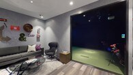 This sport simulation room offers an impressive blend of high-tech entertainment and sophisticated interior design. At the heart of the space is a full golf simulator bay, enclosed within a darkened enclosure fitted with a green artificial turf hitting area, providing an immersive and realistic gameplay experience. Recessed ceiling lighting illuminates the room with a warm, focused glow, accentuating the textured grey wall panels that lend a refined, contemporary aesthetic throughout.

Adjacent to the simulator bay, a comfortable lounge area invites guests to relax and socialize. The seating arrangement features a sleek grey sofa accented with vibrant purple throw pillows, paired with a sculptural dark wingback chair and a warm-toned metallic side table. A round glass coffee table completes the setting, adding a touch of modern elegance. The wood-look plank flooring grounds the space with a natural warmth that contrasts nicely with the cool grey tones of the walls.

Playful decorative signage, including a lit "OPEN" sign and vintage-style wall art, adds personality and a fun, lounge-like atmosphere to the room. Recessed spotlights throughout ensure the entire space is well-lit and visually inviting. This simulation room is thoughtfully designed to serve both as a serious sport training environment and an entertaining social hub.
