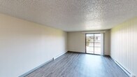 Located at 7007 N Nevada St in Spokane, WA, this two-bedroom, one-bathroom unit offers a practical and well-organized single-floor layout. The open-concept kitchen and living area form the heart of the home, where a peninsula counter with a granite-look surface and dark wood cabinetry above separates the kitchen from the living space. The kitchen is equipped with a white refrigerator, electric range, and a stainless steel sink, with ample upper and lower cabinet storage finished in warm wood tones. The living room features wood-style vinyl plank flooring, a textured ceiling, and a baseboard heating unit along one wall. A sliding glass door fills the room with natural light and opens to a private balcony with outdoor views of the surrounding landscape. Both bedrooms are carpeted and include closets with sliding panel doors, offering dedicated storage. The bathroom features a full tub and shower combination with white tile surround and chrome fixtures. Throughout the unit, neutral wall tones and consistent finishes create a cohesive and functional living environment suited for everyday comfort.