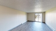 Located at 7007 N Nevada St in Spokane, WA, this two-bedroom, one-bathroom unit offers a practical and well-organized single-floor layout. The open-concept kitchen and living area form the heart of the home, where a peninsula counter with a granite-look surface and dark wood cabinetry above separates the kitchen from the living space. The kitchen is equipped with a white refrigerator, electric range, and a stainless steel sink, with ample upper and lower cabinet storage finished in warm wood tones. The living room features wood-style vinyl plank flooring, a textured ceiling, and a baseboard heating unit along one wall. A sliding glass door fills the room with natural light and opens to a private balcony with outdoor views of the surrounding landscape. Both bedrooms are carpeted and include closets with sliding panel doors, offering dedicated storage. The bathroom features a full tub and shower combination with white tile surround and chrome fixtures. Throughout the unit, neutral wall tones and consistent finishes create a cohesive and functional living environment suited for everyday comfort.