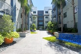 Located at 1785 Almaden Road in San Jose, California, this thoughtfully designed outdoor common area serves as a dynamic communal hub set within a modern multi-story residential community. The centerpiece of the space is a wide wood-plank promenade flanked by towering palm trees, lush greenery, and vibrant potted plants in bold orange and green vessels, creating a resort-like atmosphere under the open California sky. String lights overhead add an inviting ambiance that carries from day into evening. The promenade connects multiple distinct outdoor zones, including a spacious tiled patio area featuring a striking stone-clad fireplace and café-style seating, ideal for relaxed gatherings. A separate courtyard zone showcases a row of sculptural green planters set against the clean modern facade of the building, with polished concrete tile underfoot. At one end of the complex, a generous open-air terrace extends from an indoor clubroom equipped with a pool table and modern kitchen facilities. A wooden pergola overhead shades additional outdoor dining tables, framing views of mature trees beyond. Throughout the space, decorative rock beds, water features, and manicured plantings reinforce a cohesive, contemporary aesthetic that blends urban living with an inviting outdoor lifestyle.