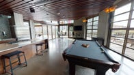 Located at 2106 5th Ave in Seattle's Belltown neighborhood, this single-floor space combines sophisticated design with versatile functionality. The interior is defined by striking rich wood slat ceilings that run throughout, polished concrete floors, and floor-to-ceiling glass walls that flood the space with natural light and frame views of the surrounding Seattle skyline.

The main gathering area centers around a full-size pool table and open lounge space, with wall-mounted flat-screen televisions positioned for easy viewing. Large glass overhead doors open directly onto a spacious outdoor terrace finished with wood decking and outdoor seating, creating a seamless indoor-outdoor flow. Sculptural pendant light fixtures add warmth and visual interest throughout.

The fully equipped chef's kitchen features rich walnut cabinetry, mosaic tile backsplash, granite countertops, a large center island with gas cooktop, stainless steel double wall ovens, a full-size refrigerator, and additional drawer refrigeration units — well-suited for catering or event use.

Throughout the space, the interplay of warm wood tones, polished concrete, and expansive glazing creates a contemporary yet inviting ambiance ideal for gatherings, events, or community use.