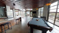 Located at 2106 5th Ave in Seattle's Belltown neighborhood, this single-floor space combines sophisticated design with versatile functionality. The interior is defined by striking rich wood slat ceilings that run throughout, polished concrete floors, and floor-to-ceiling glass walls that flood the space with natural light and frame views of the surrounding Seattle skyline.

The main gathering area centers around a full-size pool table and open lounge space, with wall-mounted flat-screen televisions positioned for easy viewing. Large glass overhead doors open directly onto a spacious outdoor terrace finished with wood decking and outdoor seating, creating a seamless indoor-outdoor flow. Sculptural pendant light fixtures add warmth and visual interest throughout.

The fully equipped chef's kitchen features rich walnut cabinetry, mosaic tile backsplash, granite countertops, a large center island with gas cooktop, stainless steel double wall ovens, a full-size refrigerator, and additional drawer refrigeration units — well-suited for catering or event use.

Throughout the space, the interplay of warm wood tones, polished concrete, and expansive glazing creates a contemporary yet inviting ambiance ideal for gatherings, events, or community use.
