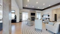 This single-floor space is a thoughtfully designed communal lounge that combines modern aesthetics with inviting comfort. The open-plan layout is anchored by a sleek fireplace flanked by large windows dressed in sheer white curtains, allowing generous natural light to fill the room. A wall-mounted flat-screen television above the fireplace serves as a central focal point, making the space equally suited for relaxed socializing or entertainment.

The seating area features a carefully curated arrangement of upholstered armchairs and accent chairs in neutral tones, complemented by a bold chevron-patterned chair that adds visual interest. A striped area rug grounds the seating arrangement atop light-toned hardwood flooring, contributing to the room's warm, contemporary feel.

Elevated ceiling details with recessed lighting and a contrasting dark tray ceiling border add architectural dimension to the space. Along one side, a dark-cabineted refreshment or service station provides functional convenience. Decorative shelving units display lush greenery and curated accessories, lending the space a polished yet welcoming atmosphere. The overall design balances clean lines with soft furnishings, creating an environment that is both visually refined and practically comfortable.