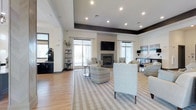This single-floor space is a thoughtfully designed communal lounge that combines modern aesthetics with inviting comfort. The open-plan layout is anchored by a sleek fireplace flanked by large windows dressed in sheer white curtains, allowing generous natural light to fill the room. A wall-mounted flat-screen television above the fireplace serves as a central focal point, making the space equally suited for relaxed socializing or entertainment.

The seating area features a carefully curated arrangement of upholstered armchairs and accent chairs in neutral tones, complemented by a bold chevron-patterned chair that adds visual interest. A striped area rug grounds the seating arrangement atop light-toned hardwood flooring, contributing to the room's warm, contemporary feel.

Elevated ceiling details with recessed lighting and a contrasting dark tray ceiling border add architectural dimension to the space. Along one side, a dark-cabineted refreshment or service station provides functional convenience. Decorative shelving units display lush greenery and curated accessories, lending the space a polished yet welcoming atmosphere. The overall design balances clean lines with soft furnishings, creating an environment that is both visually refined and practically comfortable.