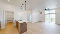 This well-designed single-floor unit offers a thoughtful layout that maximizes functionality within a compact footprint. The open-plan kitchen and living area serves as the heart of the space, anchored by a contemporary kitchen island with a white quartz countertop, undermount sink, and wood-toned cabinetry. A trio of pendant lights hangs above the island, complementing the recessed lighting throughout. Stainless steel appliances, including a refrigerator, range, and microwave, complete the kitchen. Light hardwood-style flooring flows throughout the main living areas, contributing to a bright and cohesive aesthetic reinforced by white walls and clean-lined finishes. The living area features a ceiling fan and access to an outdoor balcony, where scenic views can be enjoyed. The bedroom is carpeted for comfort, features a ceiling fan, and receives natural light through a window overlooking the outdoors. The bathroom includes a wide vanity with wood-toned cabinetry, white countertop, and large mirror. A dedicated laundry room houses a full-size washer and dryer alongside built-in shelving, adding practical storage to this efficiently designed space.