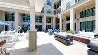 Located at 4008 Marriott Dr in Raleigh, NC, this inviting outdoor courtyard serves as a central gathering space framed by a multi-story residential building on all sides. The courtyard features a spacious stone-paved terrace that provides ample room for relaxation and socializing. Large structural columns line the covered perimeter walkways, adding an architectural elegance that complements the building's classic yet contemporary façade of neutral tones and large glass windows.

The seating arrangements throughout the courtyard are thoughtfully curated, featuring modern outdoor sofas, lounge chairs, and accent tables styled with blue and white cushions, offering a clean and sophisticated aesthetic. Colorful potted plants and flower beds add natural vibrancy to the space, softening the hardscape surroundings. A striped patio umbrella provides additional shade in one corner, enhancing comfort during sunnier days.

The open-air design allows abundant natural light to flood the courtyard, while the surrounding building's balconies and glass facades create a sense of enclosure without feeling confined. The overall ambiance is polished, welcoming, and well-suited for both quiet relaxation and casual gatherings.