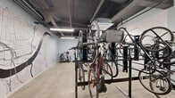Located in the heart of downtown Cincinnati at 201 W Fourth St, this dedicated bicycle storage room offers a well-organized, single-floor facility designed to accommodate a large number of bikes efficiently. The space is outfitted with multiple heavy-duty black steel vertical and horizontal bike racks, maximizing capacity while keeping the floor area navigable. A standout feature is the large-scale Cincinnati street map mural rendered in black line art on the light blue-painted walls, adding a distinctive urban character to the room. The polished concrete floors are durable and easy to maintain, suited for the practical demands of a high-use storage area. Exposed ceiling infrastructure, including conduit and ductwork painted dark, contributes to the room's industrial aesthetic. Bright linear fluorescent lighting ensures the space is well-illuminated throughout. A bike repair and inflation station is also present, providing added convenience for cyclists. Double glass-paneled doors offer secure and accessible entry and exit. Overall, the room is thoughtfully designed to serve the needs of urban cyclists, combining functional storage solutions with a visually engaging environment that reflects Cincinnati's city identity.