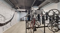 Located in the heart of downtown Cincinnati at 201 W Fourth St, this dedicated bicycle storage room offers a well-organized, single-floor facility designed to accommodate a large number of bikes efficiently. The space is outfitted with multiple heavy-duty black steel vertical and horizontal bike racks, maximizing capacity while keeping the floor area navigable. A standout feature is the large-scale Cincinnati street map mural rendered in black line art on the light blue-painted walls, adding a distinctive urban character to the room. The polished concrete floors are durable and easy to maintain, suited for the practical demands of a high-use storage area. Exposed ceiling infrastructure, including conduit and ductwork painted dark, contributes to the room's industrial aesthetic. Bright linear fluorescent lighting ensures the space is well-illuminated throughout. A bike repair and inflation station is also present, providing added convenience for cyclists. Double glass-paneled doors offer secure and accessible entry and exit. Overall, the room is thoughtfully designed to serve the needs of urban cyclists, combining functional storage solutions with a visually engaging environment that reflects Cincinnati's city identity.