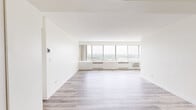 This well-appointed 2-bedroom, 1-bathroom unit offers a clean, bright aesthetic throughout, characterized by crisp white walls and a thoughtfully arranged single-floor layout. The spacious living room is a standout feature, showcasing a wide bank of floor-to-ceiling windows that flood the space with natural light and frame sweeping panoramic views of the surrounding cityscape and greenery. Light wood-style plank flooring extends throughout the living and hallway areas, lending a modern, cohesive feel.

The generously sized primary bedroom features plush carpet flooring, large windows with elevated outdoor views, and a wide louvered bi-fold closet providing ample storage. The second bedroom mirrors a similar layout, also carpeted and outfitted with a louvered closet. Both rooms maintain the unit's consistent bright and airy character.

The kitchen is efficiently designed with white cabinetry, granite-toned countertops, and white appliances including a range, microwave, and refrigerator, all set on wood-style flooring. The full bathroom includes a tiled tub-shower combination, a vanity with warm-toned countertop, white cabinetry, and a mirrored medicine cabinet above. A connecting hallway ties all rooms together, completing this well-organized and light-filled residence.