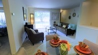 This single-floor residence offers a bright and inviting layout designed for comfortable everyday living. The combined living and dining area forms the heart of the space, bathed in natural light streaming through a large sliding glass door adorned with vertical blinds. The door opens to a view of lush greenery outside, creating a seamless connection between the interior and the outdoors. The living area is generously proportioned, accommodating two plush sofas arranged around a glass-top coffee table, all set atop a soft area rug over light-toned carpeting. Decorative wall art and accent mirrors add personality and visual interest to the neutral-toned walls. The dining area sits adjacent to the living space, featuring a round glass dining table set with colorful tableware and a vibrant floral centerpiece that adds a cheerful pop of color. The open-concept flow between the dining and living zones makes the space feel expansive and well-connected, ideal for both relaxing and entertaining. The overall ambiance is warm and welcoming, with a neutral color palette complemented by thoughtful décor accents throughout.