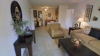 This single-floor residence offers a comfortable and functional layout centered around an open-concept living, dining, and kitchen area. The living room is carpeted in a neutral tone and furnished with a warm, inviting arrangement, including plush seating and dark wood accent tables. A ceiling fan overhead provides both light and air circulation, adding a practical touch to the space. The room opens naturally into a casual dining area, defined by dark wood furnishings and framed wall art, creating a cohesive and intimate setting for meals. Beyond the dining area, the kitchen features warm wood cabinetry, tile flooring, and standard appliances including a refrigerator and range, offering a well-equipped cooking space. A peninsula-style counter connects the kitchen to the living and dining zones, enhancing the open flow and making the layout ideal for everyday living and entertaining. The consistent use of neutral beige and warm brown tones throughout creates a unified, calming ambiance. The space is practical and well-proportioned, with each area clearly defined yet visually connected, making it feel open and easy to navigate.