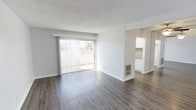 This single-floor space offers a clean, minimalist aesthetic defined by neutral grey-toned walls, a textured white ceiling, and wide-plank hardwood-style flooring that runs throughout. The main living area is generously proportioned, bathed in natural light streaming through a large sliding glass door fitted with vertical blinds. The door opens to an outdoor space, providing a seamless indoor-outdoor connection and adding a sense of openness to the interior. A wall-mounted heating unit is integrated neatly into the living area, maintaining the streamlined look of the space. The open layout flows naturally toward an adjoining kitchen, visible through a wide doorway. The kitchen features white cabinetry and a tiled backsplash, maintaining the bright, cohesive color palette established throughout. The overall ambiance is light, airy, and move-in ready, with a practical, uncluttered design that offers flexibility for a variety of furniture arrangements and personal styles.