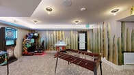Located in Scottsdale, Arizona, this well-appointed amenity space delivers an impressive entertainment experience across a single thoughtfully designed floor. The game room serves as the centerpiece, featuring patterned carpeting and a striking floor-to-ceiling saguaro cactus mural that pays homage to the Sonoran Desert landscape, lending the space a distinctly Southwest character.

The main gaming area is equipped with a foosball table, an arcade racing game, and an air hockey table, offering a variety of interactive entertainment. A dedicated alcove houses a classic 007-themed pinball machine alongside an electronic BullShooter dartboard, providing additional options for guests. Recessed tray ceilings with accent lighting add architectural depth and a polished finish throughout.

A lounge area adjacent to the game room features a bistro-style high-top table with bar seating, complemented by framed abstract artwork on the walls — creating an inviting spot to relax between games.

A separate, fully enclosed indoor golf simulator room rounds out the offerings, complete with a projected course display, professional-grade turf hitting mats, overhead projector, and launch monitor technology — ideal for golfers of all skill levels. The overall ambiance is energetic yet refined, blending recreational functionality with stylish, regionally inspired design elements.