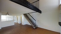This two-story space makes a strong architectural statement with its open, loft-inspired design. The main living area is anchored by warm hardwood flooring that extends throughout, complemented by clean white walls that enhance the sense of light and space. A dramatic open-tread staircase with a sleek black metal railing serves as a functional centerpiece, connecting the lower level to the upper floor while adding an unmistakably industrial character to the interior. The double-height ceiling above the main living area amplifies the sense of volume and openness, a standout feature that sets the space apart. The upper level is bordered by a matching black metal railing, offering a visual continuity that ties the two floors together cohesively. Toward the rear of the main floor, a glimpse into the kitchen area reveals a practical layout with cabinetry and countertop space, alongside what appears to be a corridor leading to additional rooms. The overall ambiance blends industrial and modern elements — raw metal details contrast with the warmth of the wood floors and the brightness of the white walls — resulting in a space that feels both stylish and functional.