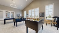 Located at 3400 Club Way Ct in Powell, Ohio, this community amenity center offers a thoughtfully designed, single-floor layout spanning a generously sized footprint. The space is organized into distinct functional areas that cater to recreation, socializing, and fitness.

The standout game room features soaring vaulted ceilings and is bathed in natural light from large windows with white blinds. Dramatic circular pendant light fixtures hang overhead, illuminating a pool table, two foosball tables, and bar-height seating — all set on a neutral patterned carpet that adds subtle texture underfoot.

Adjacent to the game room, a bright lounge and meeting area provides comfortable seating with upholstered curved chairs arranged around round and rectangular tables. Large windows frame outdoor views, and a wall-mounted TV adds a functional touch. The area connects seamlessly to a stylish bar or refreshment station, complete with a decorative art-deco inspired counter facade, a light quartz countertop, pendant lighting, and a built-in beverage refrigerator framed by sleek subway tile.

A glazed interior wall and French doors reveal a well-equipped fitness center featuring cardio machines and strength training equipment, bathed in natural light. Throughout, the space maintains a cohesive warm gray palette, white trim, and curated artwork, creating a polished and welcoming community environment.