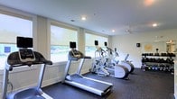 Located at 1109 Matteo Dr in Wilmington, North Carolina, this single-floor fitness center is designed to provide a functional and comfortable workout environment. The space is equipped with a selection of cardio machines, including treadmills, an elliptical trainer, and a stair climber, each outfitted with individual display screens for an enhanced exercise experience. Large windows line one wall of the room, allowing generous amounts of natural light to fill the space while offering pleasant views of the surrounding greenery outdoors. The bright, neutral-toned walls and recessed ceiling lighting contribute to a clean, open atmosphere. A ceiling fan is mounted overhead, providing consistent airflow throughout the room to keep the environment comfortable during workouts. The floor is finished with durable, dark rubber gym flooring, which is both practical and easy to maintain. The layout is well-organized, allowing for easy movement between equipment. Overall, this fitness center offers a well-lit, properly ventilated, and thoughtfully equipped space suitable for a variety of cardio-focused exercise routines.