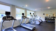 Located at 1109 Matteo Dr in Wilmington, North Carolina, this single-floor fitness center is designed to provide a functional and comfortable workout environment. The space is equipped with a selection of cardio machines, including treadmills, an elliptical trainer, and a stair climber, each outfitted with individual display screens for an enhanced exercise experience. Large windows line one wall of the room, allowing generous amounts of natural light to fill the space while offering pleasant views of the surrounding greenery outdoors. The bright, neutral-toned walls and recessed ceiling lighting contribute to a clean, open atmosphere. A ceiling fan is mounted overhead, providing consistent airflow throughout the room to keep the environment comfortable during workouts. The floor is finished with durable, dark rubber gym flooring, which is both practical and easy to maintain. The layout is well-organized, allowing for easy movement between equipment. Overall, this fitness center offers a well-lit, properly ventilated, and thoughtfully equipped space suitable for a variety of cardio-focused exercise routines.