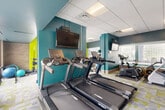 Located at 401 Cumberland Ave in Portland, Maine, this single-floor amenity facility offers a comprehensive wellness experience across a spacious footprint. The space is anchored by a large indoor swimming pool, surrounded by a tiled pool deck, structural columns, and expansive glass block windows that diffuse natural light throughout the area, creating a bright and tranquil atmosphere. Seating areas along the poolside provide a comfortable space for relaxation.

Adjacent to the pool, the fitness center is vibrant and energetic, featuring bold teal and lime green accent walls paired with patterned, multi-tone carpet tiles. The gym is well-equipped with cardio machines including StairMasters, ellipticals, stationary bikes, and treadmills, as well as strength training equipment such as a squat rack, free weights, and stability balls. Large windows overlooking the pool area keep the workout space open and connected.

A welcoming reception or lounge area features a striking wood-panel accent wall, terrazzo flooring, and mid-century modern décor including a starburst wall clock and tall potted plants, lending the space a timeless, polished character. The kitchen is functional and well-appointed, with white cabinetry, light countertops, stainless steel appliances including a refrigerator, dishwasher, and microwave, and wood-look flooring. Drop ceilings with recessed and panel lighting are consistent throughout, giving the entire facility a clean, cohesive finish.