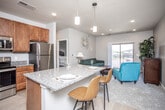 This single-floor residence offers a thoughtfully designed open-concept layout that seamlessly connects the kitchen, dining, and living areas. The kitchen is a functional centerpiece, featuring rich warm-toned cabinetry extending to the ceiling for ample storage, granite countertops, stainless steel appliances including a refrigerator, range, and microwave, and a peninsula island with bar seating for two—illuminated by pendant lighting overhead. Light wood-style flooring runs throughout the kitchen and entry areas, while recessed lighting keeps the space bright and welcoming.

The living area transitions naturally from the kitchen, finished with plush carpeting and a sliding glass door that opens to a private balcony with exterior views. The bedroom is generously sized, featuring soft carpet, a ceiling fan with light fixture, and windows that bring in natural light. It is accessed directly from the main living space for convenient flow.

The bathroom is clean and well-equipped, offering a granite-topped vanity with warm wood cabinetry, a combination tub and shower, and neutral gray walls that complement the overall palette. Throughout the unit, a consistent color scheme of soft grays and warm wood tones creates a cohesive, comfortable atmosphere.