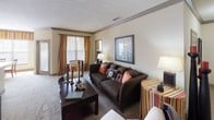 This single-story residence offers a thoughtfully designed layout with warm, neutral tones and consistent finishes throughout. The living room is generously proportioned, featuring crown molding, a textured ceiling, and plush carpeting. Large windows dressed in striped drapery allow abundant natural light to fill the space, which comfortably accommodates a full seating arrangement alongside a substantial coffee table and decorative shelving.

Adjacent to the living room, the dining area features tile flooring and a pendant light fixture above a glass-topped table, with a wide window providing an open, airy feel. The kitchen offers rich wood cabinetry, dark countertops, tile flooring, and black appliances including a refrigerator, dishwasher, and range.

The primary bedroom is carpeted and well-lit, with dual windows, ceiling fan, and ample dresser space. An en-suite bathroom includes a wood-cabinet vanity, tile flooring, a large mirror, and access to a spacious walk-in closet with built-in shelving. Additional bedrooms are carpeted and bright, each with generous window coverage and tasteful wall accents, completing a comfortable and cohesive living environment.
