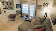 This single-floor space is thoughtfully designed to maximize comfort and functionality. The living area serves as the heart of the space, centered around a plush L-shaped sectional sofa accented with decorative throw pillows in warm earth tones. A sleek round glass-top coffee table sits at the center, adding a touch of modern elegance to the room. The flooring is covered in neutral-toned carpet, contributing to the warm and inviting ambiance throughout.

Natural light flows generously into the space through large windows fitted with horizontal blinds, flanking a glass-paneled door that opens to an outdoor balcony — offering a seamless connection between the interior and exterior. A wall-mounted flat-screen television is positioned above a set of floating shelves, which provide practical storage for books, décor, and personal items. Decorative wall art adds personality to the room, with a distinctive circular wall display offering a stylish focal point.

A woven accent chair provides additional seating and a casual, relaxed character to the living area. The overall aesthetic is contemporary and understated, with neutral wall tones complementing the furnishings. The layout is efficient yet comfortable, making excellent use of the available space to create a welcoming and well-appointed environment.