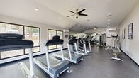 Located at 500 Brooksboro Terr in Nashville, TN, this fitness center offers a well-appointed, single-room workout facility designed to support a comprehensive exercise routine. The space features vaulted ceilings that lend an open, airy feel, complemented by recessed lighting and ceiling fans that maintain a comfortable environment during workouts. Large windows along the perimeter flood the room with natural light and provide views of the surrounding greenery, creating an inviting atmosphere. The flooring throughout is a durable dark rubber material, ideal for high-traffic fitness use. A row of treadmills and elliptical trainers lines one side of the room, providing ample cardio options, while the opposite area is dedicated to strength training, featuring a multi-station cable machine, a weight bench, and stationary bikes. Full-wall mirrors on one side of the room visually expand the space and allow users to monitor their form. A wall-mounted split air conditioning unit ensures climate control year-round. The facility is accessible via a set of glass double doors and is equipped with safety essentials including a fire extinguisher and clearly marked exit signage, making it a functional and well-maintained fitness amenity.