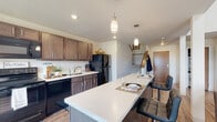 This two-story residence is defined by its dramatic double-height ceilings in the main living area, which create an expansive, airy atmosphere flooded with natural light from a clerestory window positioned high on the wall. The living room opens directly to a private balcony through sliding glass doors, extending the living space outdoors and providing views of the surrounding area. Warm hardwood-style flooring runs throughout the main level, complementing the neutral color palette of white and light gray walls.

The kitchen is a functional centerpiece, featuring rich dark-stained cabinetry extending to the ceiling, light quartz countertops, black appliances including a range, microwave, dishwasher, and refrigerator, and a generously sized island with bar seating and pendant lighting overhead. A staircase near the kitchen entry connects the two floors. The dining area sits adjacent to the kitchen, benefiting from a large window with neighborhood views.

Bedrooms are located on the upper level, offering carpeted or hardwood flooring and sizable windows that bring in natural light. Each bathroom is finished with dark wood vanity cabinetry, white countertops, hardwood-style flooring, and glass-enclosed showers, maintaining a consistent, modern aesthetic throughout the residence.