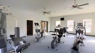 This single-story fitness facility offers a functional and comfortable workout environment designed to accommodate a variety of exercise needs. The spacious room is finished with neutral grey walls and grey carpeted flooring, creating a clean, understated aesthetic throughout. Natural light filters in through multiple windows along the perimeter, brightening the space and offering views of the surrounding exterior. Two ceiling fans are mounted overhead, promoting air circulation and comfort during workouts.

The room is equipped with a selection of cardio machines, including elliptical trainers, a stationary recumbent bike, and a treadmill, providing users with diverse cardiovascular training options. Additional equipment, including what appears to be a leg curl bench, is also present. A wall-mounted flat-screen television is positioned at a convenient viewing height, adding an entertainment element for users during exercise sessions.

A doorway on one side of the room leads to an adjacent space, suggesting connectivity to other amenity areas. Glass doors near the back of the room provide access to an outdoor area, further enhancing the sense of openness. Overall, the space is practical, well-lit, and thoughtfully arranged for fitness use.