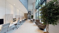 Located at 63 E Lake Street in the heart of Chicago's Loop, this sophisticated ground-floor amenity space exemplifies contemporary commercial design. The open layout is anchored by dramatically soaring double-height ceilings and expansive floor-to-ceiling glass curtain walls that flood the interior with natural light and frame sweeping views of the surrounding urban streetscape and landscaped outdoor areas.

Polished large-format white tile flooring runs throughout, lending a clean, cohesive aesthetic to the entire space. Textured sculptural wall panels in varying shades of gray add architectural depth and visual interest, complementing the neutral palette. Oversized indoor trees and curated greenery are positioned throughout, softening the modern design and bringing a natural, organic element indoors.

A dedicated workstation area along one wall offers built-in desk surfaces with ergonomic seating, ideal for focused work or quick tasks. Adjacent lounge zones feature plush leather swivel chairs and modular seating arranged around low tables, with pendant globe chandeliers overhead adding an elegant finishing touch. Dual wall-mounted flat-screen televisions anchor a secondary lounge area, creating a comfortable media-ready gathering space.

The kitchenette is outfitted with sleek matte cabinetry, white quartz countertops, a built-in cooktop, stainless steel sink, beverage station, and open shelving — providing full functionality within a refined, minimalist aesthetic.