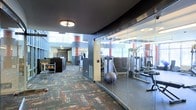 This single-floor fitness facility is designed with both functionality and modern aesthetics in mind. The space is divided into distinct zones, most notably a glass-enclosed gym area that offers full visibility into the equipment room, which houses strength training machines, free weights, benches, and stability balls. This transparent enclosure creates a sense of openness while maintaining a dedicated workout environment.

The surrounding corridor and common areas feature bold design elements, including striking terracotta-red cylindrical columns that provide strong visual contrast against the dark ceiling and gray walls. The flooring throughout the common area consists of richly patterned carpet in earthy tones of rust, brown, and gray, adding warmth and texture to the space.

Natural light filters in through large windows and elevated skylights, brightening the interior and contributing to an open, airy atmosphere. Recessed and flush-mount ceiling fixtures provide consistent artificial lighting throughout. A reception or service counter is visible near the entrance, suggesting a managed, amenity-style facility. Overall, the space combines professional fitness functionality with a polished, contemporary design.