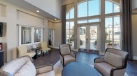 This single-floor community clubhouse is designed to impress from the moment you enter. The grand reception area greets visitors with soaring double-height ceilings and an expansive wall of floor-to-ceiling arched windows that flood the space with natural light and frame views of the surrounding landscaped grounds. French glass doors at the entrance enhance the open, airy feel throughout.

The main lounge area is anchored by a tiled fireplace with a classic white mantel, creating a warm and inviting focal point. Comfortable seating arrangements — including upholstered chairs and sofas in neutral tones — are thoughtfully positioned to encourage conversation and relaxation. A wall-mounted flat-screen television adds a modern touch to the living area.

A dedicated coffee and refreshment station, housed in a sleek dark cabinet, provides a convenient amenity for residents. Adjacent to the lounge, glass-paneled doors lead to a game room featuring a pool table, ideal for recreation and socializing. Additional flex spaces are accessible via interior French doors, offering versatility for a range of uses.

Dark tile flooring runs throughout, providing a rich, durable foundation that complements the neutral gray and white color palette. Recessed lighting, decorative artwork, and carefully selected furnishings complete this polished and functional community gathering space.