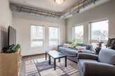 This single-floor residential unit offers a thoughtfully laid-out living space with a blend of modern finishes and industrial design elements. The open-concept living and dining area is a central highlight, featuring exposed ductwork that adds an urban, loft-inspired character. Multiple windows flood the space with natural light and provide views of the surrounding neighborhood. A sliding glass door in the dining area leads to a private balcony, extending the living space outdoors.

The kitchen is well-equipped with dark espresso cabinetry, granite countertops, a breakfast bar with pendant lighting, and full appliances including a refrigerator, stove, and microwave. Wood-look flooring runs throughout the main living areas, adding warmth and cohesion.

The unit includes two bedrooms, each with carpeted floors, closet storage, and windows that bring in natural light. A narrow hallway connects the private areas of the unit to the main living spaces, lined with smooth white walls and wood-look flooring that continues from the main area. The bathroom, visible at the end of the hallway, is clean and functional. Overall, the unit presents a practical and comfortable layout with a distinctive industrial-modern aesthetic.