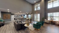 This elegantly designed single-floor clubhouse offers a seamless blend of modern sophistication and functional communal living. The grand main lounge commands attention with its dramatically soaring double-height ceilings, warm hardwood-style flooring, and a curated mix of contemporary furnishings, including plush seating and bold emerald green velvet accent chairs accented with gold bases. Statement pendant lighting in matte black cascades from above, complementing the space's refined color palette of deep grays and charcoal tones. Expansive windows and French doors flood the lounge with natural light and provide views of the surrounding greenery, creating an inviting, airy atmosphere. A dedicated seating nook features a round wood table flanked by neutral linen chairs, ideal for smaller gatherings or quiet conversation. The catering kitchen is a standout feature, showcasing an elongated granite-topped island with bar seating, dark espresso cabinetry, decorative lattice pendant lights, stainless steel appliances, and mirrored backsplash panels that amplify depth and light. The entry corridor sets an impressive tone with its soaring walls, curated artwork, and stylish console furnishings. Throughout, the space maintains a cohesive, upscale aesthetic well-suited for both social events and everyday community use.