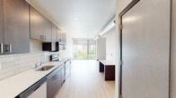 Located in Iowa City, IA, this contemporary studio offers a well-designed single-floor layout that maximizes every square foot. The open living area is anchored by large floor-to-ceiling windows that flood the space with natural light and offer views of the surrounding urban landscape and greenery. Light-toned hardwood-style flooring runs throughout, complementing the clean, minimalist aesthetic.

The galley-style kitchen is thoughtfully appointed with rich dark wood cabinetry extending to the ceiling for ample storage, crisp white quartz countertops, and a classic subway tile backsplash. Stainless steel appliances — including a refrigerator, electric range, built-in microwave, and dishwasher — complete the functional and stylish setup. An exposed ventilation duct adds an industrial accent to the living area.

The bathroom features a glass-enclosed shower finished with large-format stone-look tile in warm gray tones, lending a spa-like quality to the space. A built-in shelf within the shower provides convenient storage. The overall design of the unit reflects a modern urban sensibility, combining clean lines, quality finishes, and efficient use of space to create a comfortable and cohesive living environment.