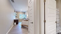 Located at 6620 Chestnut St in Lino Lakes, Minnesota, this one-bedroom suite offers a thoughtfully designed single-floor layout that maximizes functionality within a compact footprint. Light wood-style luxury vinyl flooring runs throughout the space, creating a clean and cohesive aesthetic.

The spacious bedroom serves as the heart of the suite, accommodating a full-size bed with room to spare. Recessed lighting and large windows allow natural light to fill the room, while neutral warm-toned walls contribute to a calm and welcoming ambiance. A small built-in countertop area with dark cabinetry and a marble-style surface provides a convenient workspace or kitchenette station. Open wall shelving adds additional storage and display options.

The entrance hallway connects seamlessly to the main living area and features an arched full-length mirror, adding depth and elegance to the space.

The bathroom is designed with accessibility in mind, equipped with a wall-mounted sink, a large mirror with overhead lighting, grab bars near the toilet, and a shower area with a curtain enclosure. The consistent flooring and neutral palette carry throughout, reinforcing the suite's cohesive modern design.

Overall, this suite presents a practical yet stylish living environment, ideal for those seeking comfortable, accessible, and well-finished accommodations in Lino Lakes.