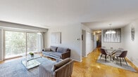 Located at 6900 Marsue Dr in Baltimore, MD, this single-floor residence offers a thoughtfully arranged layout with a cohesive, modern aesthetic throughout. Warm parquet hardwood flooring runs continuously across the main living areas, lending a classic yet inviting character to the space.

The living room is generously proportioned, bathed in natural light through a large sliding glass door that opens to an outdoor view of mature trees, creating a seamless connection between interior and exterior. A built-in media wall adds a functional design element alongside the comfortable seating area. The adjoining dining area features a decorative chandelier overhead and flows naturally from the living space, making the open-plan arrangement ideal for both daily living and entertaining.

The kitchen is efficiently designed with rich wood cabinetry, light countertops, a gas range, built-in microwave, and a window that brings in additional natural light. Two bedrooms offer comfortable accommodations, each with parquet flooring and window access to outdoor greenery. A well-organized walk-in closet with built-in shelving provides notable storage. The bathroom features white subway tile wainscoting, a wood-toned vanity, and clean, neutral finishes throughout.