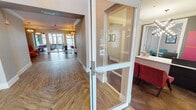 Linden Greeneway's amenity center is a thoughtfully designed, single-floor facility that blends contemporary aesthetics with functional community spaces. The entrance greets visitors through expansive floor-to-ceiling glass doors, opening into a welcoming corridor adorned with herringbone-patterned hardwood-style flooring that flows throughout the common areas.

The leasing and reception area features a sleek, dark wood desk set against a bold magenta and charcoal accent wall, illuminated by a striking starburst chandelier with exposed bulbs. An adjacent private office provides a quiet space for consultations.

The open-plan lounge is anchored by a dramatic stone-clad accent wall housing a wall-mounted television and a linear fireplace below. Soaring double-height ceilings and large windows flood the space with natural light, while pendant lighting adds warmth. The communal kitchen is outfitted with gray shaker cabinetry, a mosaic tile backsplash, stainless steel appliances, and a wide island with bar seating beneath a corrugated metal ceiling detail and pendant lights.

The fitness center is a vibrant, well-equipped space featuring multi-station cable machines, treadmills, free weights, and large mirrors, all set atop colorful striped flooring with ample natural light from oversized windows.