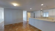 This single-floor space showcases a practical, open-concept layout that seamlessly connects the main living area to the kitchen. The flooring throughout is a warm terracotta tile, lending a classic, earthy character to the interior and providing a durable, easy-to-maintain surface.

The kitchen is defined by a raised peninsula with a light-colored countertop, offering both workspace and a natural divider between the cooking and living areas. Upper cabinetry in white finishes is visible on the far wall, contributing to a clean, functional aesthetic. A utility sink area is also noted along the kitchen's perimeter.

The living area is bright and open, with white-painted walls that reflect light from a combination of flush-mount ceiling fixtures and recessed lighting, ensuring consistent illumination throughout. The layout allows for flexible furniture arrangement within the main space.

A hallway visible in the background leads to additional rooms, including what appears to be a bathroom with a door visible at the corridor's end. The overall ambiance is neutral and move-in ready, presenting a clean canvas well-suited for personalization.