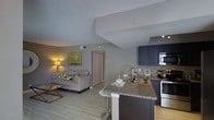 This single-floor space showcases an open-concept design that seamlessly connects the kitchen and living area, creating a cohesive and functional layout. The kitchen is a standout feature, equipped with dark cabinetry, granite countertops, and stainless steel appliances, including a range and over-the-range microwave. A peninsula with bar seating extends from the kitchen, providing additional workspace and a casual dining option. A stone tile backsplash adds texture and visual interest behind the range, complementing the overall modern aesthetic.

The living area is warm and inviting, furnished with a light-toned sofa accented by decorative pillows in muted greens and whites. A framed nature-inspired artwork adorns the wall, adding a touch of color to the neutral palette. The space benefits from light wood-style flooring throughout, contributing to a clean and airy feel. Angled ceiling elements add an architectural dimension to the interior.

A hallway visible from the main living space suggests access to additional rooms, and the overall layout is well-suited for both everyday living and entertaining.