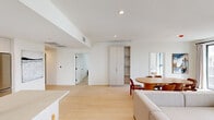 This single-floor residence offers a thoughtfully designed layout accommodating four bedrooms and three bathrooms within a generously sized floor plan. The interior embraces a clean, minimalist aesthetic defined by crisp white walls, light wood flooring, and recessed lighting throughout.

The open-concept living, dining, and kitchen area forms the heart of the home. The kitchen features white flat-front cabinetry, stainless steel appliances, a gas range, and a large island with bar seating — ideal for both daily use and entertaining. The dining area flows seamlessly into the living room, which benefits from floor-to-ceiling windows and sliding glass doors opening to a balcony with panoramic neighborhood views.

The primary bedroom provides a comfortable retreat with built-in light wood cabinetry and ample storage. Additional bedrooms maintain the same minimalist character, each with light-filled windows. The bathrooms are finished with large-format tile, black-framed glass shower enclosures, and light wood vanities with integrated sinks. Open shelving adds functional storage without visual clutter, reinforcing the cohesive, modern sensibility found throughout the entire residence.